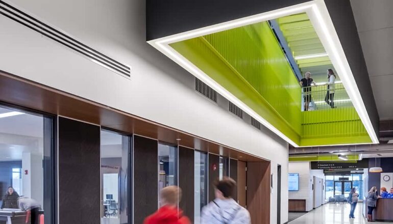 A main floor corridor leads to a welcome desk, community spaces, and a ceiling look-through into the upper level of the facility.