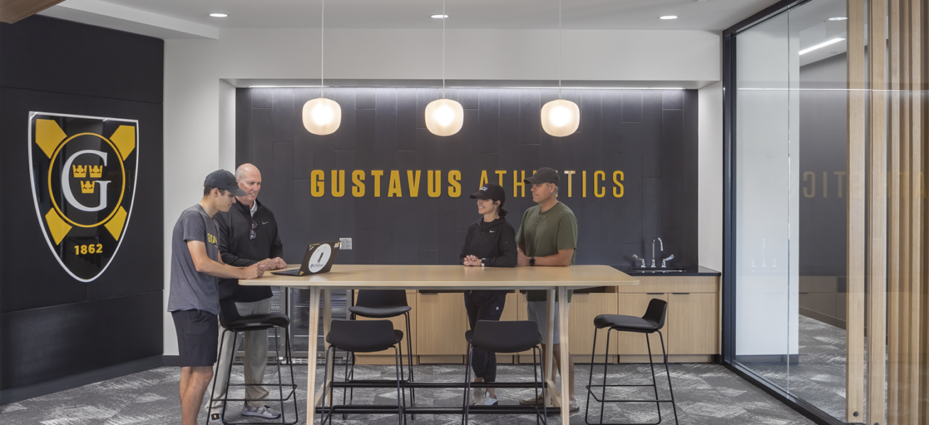 A student athlete prospect and their parents meet with a GAC coach in the recruiting room. A large Gustavus shield and branding enhance the experience