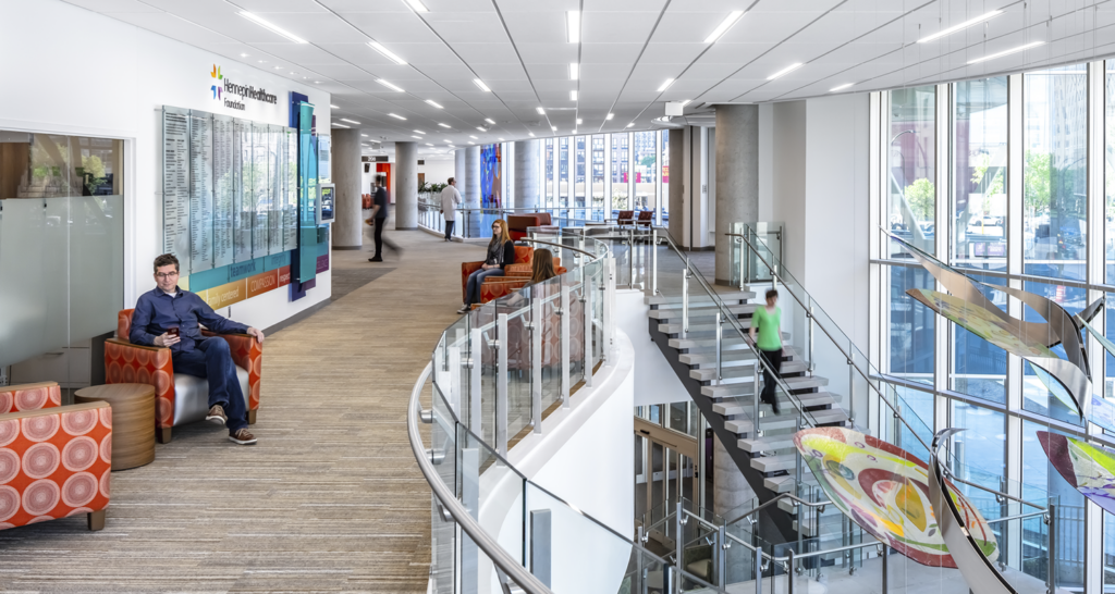 People utilize the public circulation spaces inside the Hennepin Healthcare Clinic & Specialty Center. Rainbow colored hanging artwork and a donor wall enhance the experience