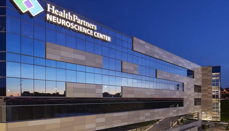 Exterior photo of the Neuroscience Center at dusk. The sunset reflects off the building's glass and the HealthPartners logo signage is lit up
