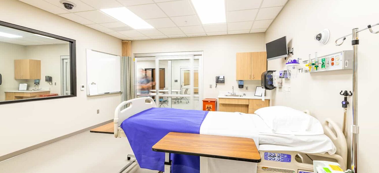 Simulation nursing exam room with hands-on learning equipment and a one-way mirror.