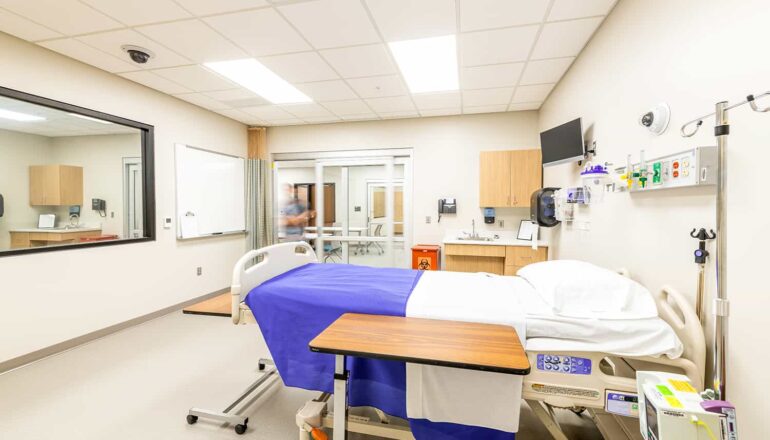 Simulation nursing exam room with hands-on learning equipment and a one-way mirror.