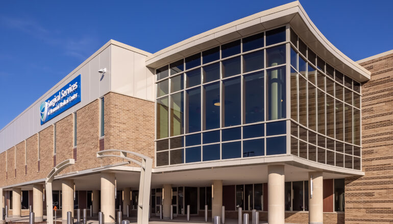 A close-up exterior view of the MMC Ashland Surgery Center. The glass-enclosed waiting room sits above the main patient drop-off and entry