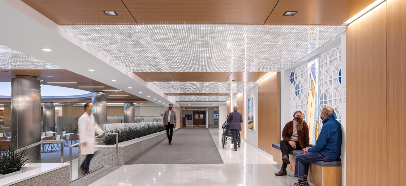 Doctors and visitors walk in and sit in a subway level corridor near the Siebens Cafeteria. Warm wood tones, enhanced lighting, detailed ceiling panels, and artwork bring joy to the space