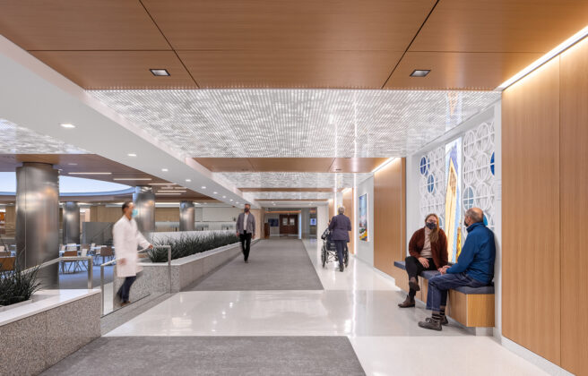 Doctors and visitors walk in and sit in a subway level corridor near the Siebens Cafeteria. Warm wood tones, enhanced lighting, detailed ceiling panels, and artwork bring joy to the space