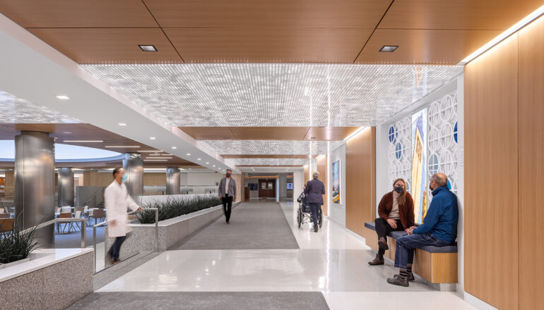 Doctors and visitors walk in and sit in a subway level corridor near the Siebens Cafeteria. Warm wood tones, enhanced lighting, detailed ceiling panels, and artwork bring joy to the space