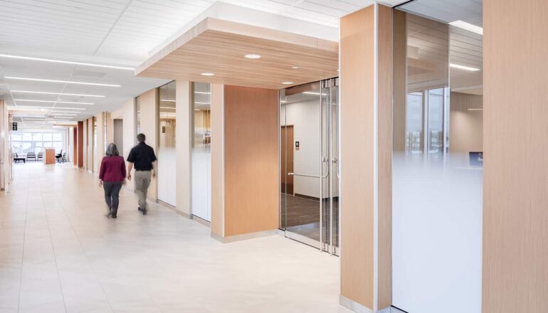 The interior circulation route with bright lighting, soft wood tones, and half opaque glass entries to program spaces.