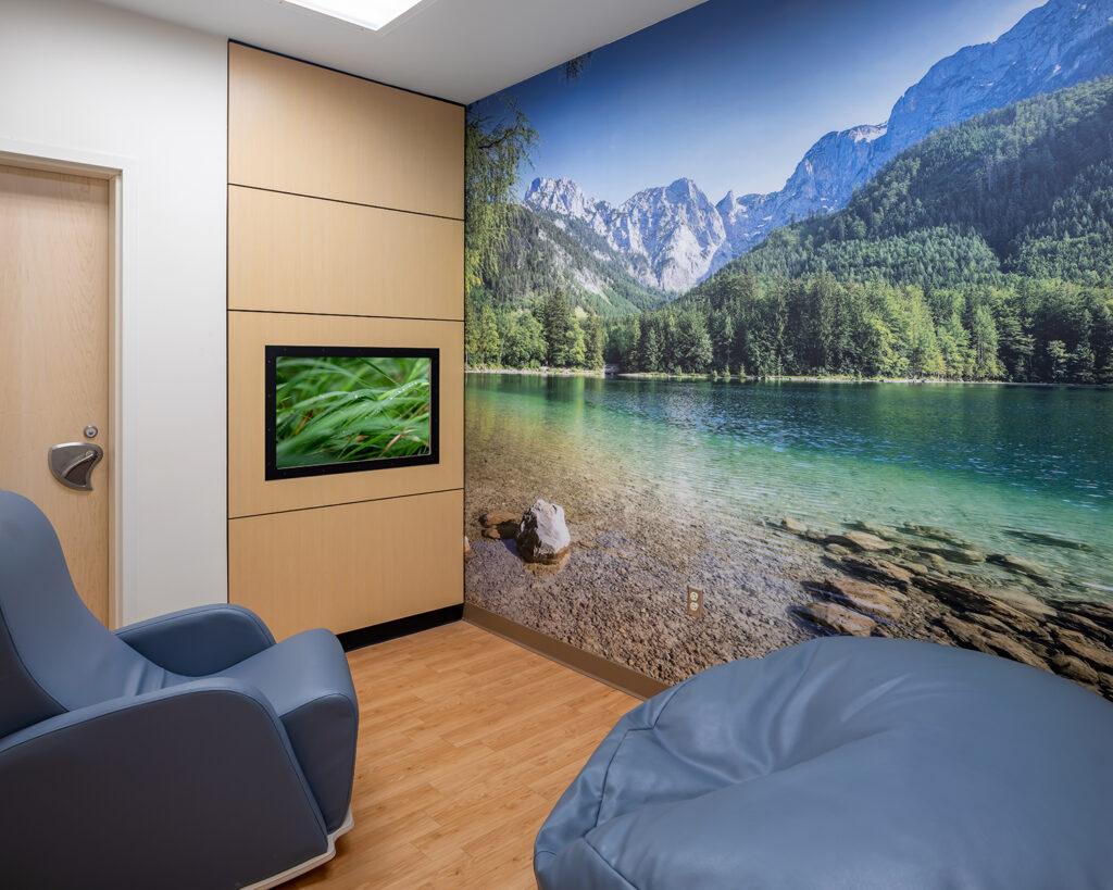 A quiet sensory room features comfy chairs, a large wall mural of a lake in the mountains, and a TV with calming videos