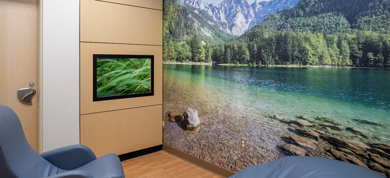 A quiet sensory room features comfy chairs, a large wall mural of a lake in the mountains, and a TV with calming videos