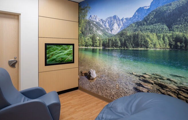 A quiet sensory room features comfy chairs, a large wall mural of a lake in the mountains, and a TV with calming videos