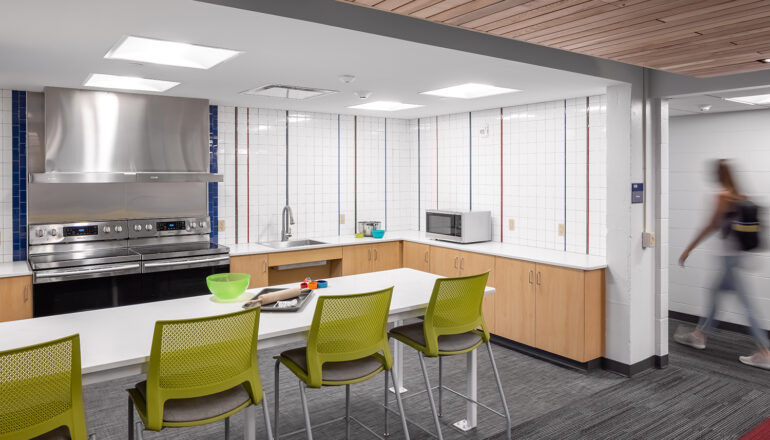 A student walks past one of the North Hall shared kitchens with neon green chairs and stainless steel appliances
