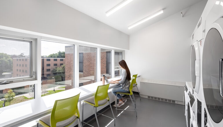 A student studies in one of the shared North Hall laundry rooms. There are neon green chairs and large windows with views to campus